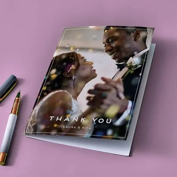 A personalised wedding thank you card featuring a photo of a couple dancing, with the text Thank You and names at the bottom, displayed on a pink surface beside two pens.